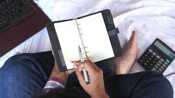 Man writing in notebook on bed with laptop and calculator video