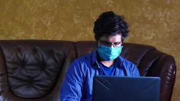 A man wearing a face mask sits on a couch with a laptop video