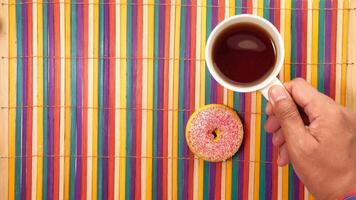 A person's hand holding a cup of tea and a donut on a colorful striped table cloth video