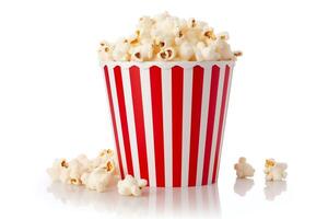 A red and white striped popcorn bucket is sitting on a white surface. The bucket is full of popped popcorn and there are some kernels spilled on the surface. photo