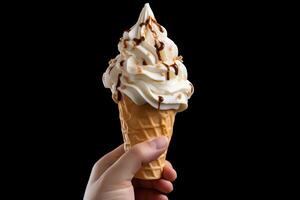 A hand holding an ice cream cone with a big swirl of soft serve vanilla ice cream topped with chocolate syrup and chopped nuts. photo