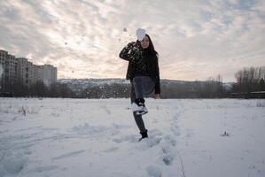 Woman standing in a snowy field, with a city skyline in the background, playfully kicking snow, enjoying a vibrant winter day. Winter fun and carefree joy in an urban setting photo