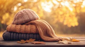 Folded sweater, scarf, and hat on a rustic wooden table, with autumn leaves in the blurred background, soft warm lighting enhancing natural textures. Autumn clothing photo
