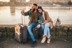 Worried couple tourists got lost on their vacation and now they are confused which way to go while sitting by the river with their luggage. photo