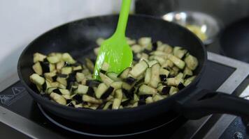 Close up view of sliced eggplant been stirred on a frying pan while been cooked. Hand stirring with green spoon in a kitchen. video