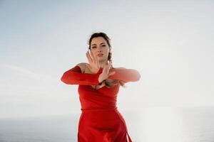 Closeup portrait of sensual young brunette woman in red dress, happily dancing outdoors isolated on blurry sea background with natural bokeh in soft warm sunset backlight. Selective focus. photo