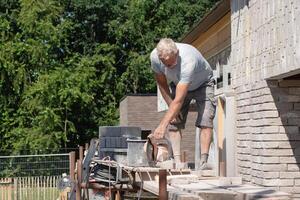 A gray-haired man saw bricks with a concrete saw of the right size for subsequent laying out in the facade of the house, High quality photo