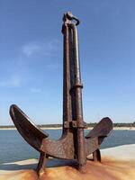A large, rusty anchor sits on a dock against a clear blue sky, symbolizing maritime history and coastal life. photo