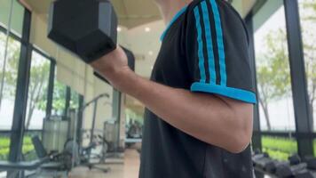 hand gripping dumbbell with a strong, determined hold. The background features a blurred view of other gym equipment, highlighting the focus on the dumbbell and the intensity of the workout, fitness video