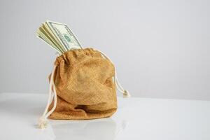 A burlap sack with a drawstring sits on a white surface with a stack of US dollar bills spilling out from the opening photo