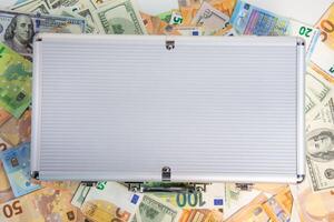 A silver briefcase is closed and lying on a white surface surrounded by US dollar bills and Euro bills photo