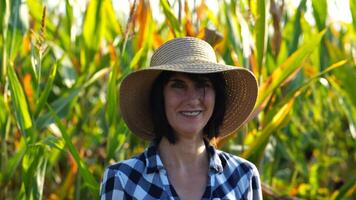 Happy smiling female farmer looks into camera standing near corn field. Portrait of adult beautiful agronomist in straw hat with maize meadow at background. Agricultural business concept. Close up photo