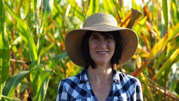 Happy smiling female farmer looks into camera standing near corn field. Portrait of adult beautiful agronomist in straw hat with maize meadow at background. Agricultural business concept. Close up photo