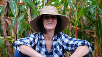 Happy smiling female farmer looks into camera sitting near corn field. Portrait of adult tired agronomist in straw hat with maize meadow at background. Concept of agricultural business. Close up photo
