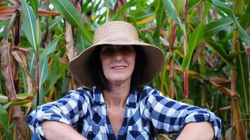 Happy smiling female farmer looks into camera sitting near corn field. Portrait of adult tired agronomist in straw hat with maize meadow at background. Concept of agricultural business. Close up photo
