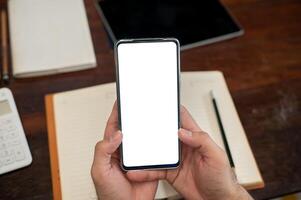 A top view of a man holding a smartphone with a white screen mockup above a desk. photo