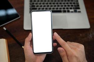 A top view of a man holding a smartphone with a white screen mockup above a desk. photo