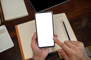 A top view of a man holding a smartphone with a white screen mockup above a desk. photo