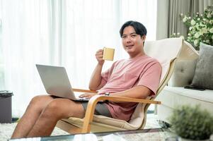 An Asian man is enjoying coffee while working on his laptop from a lounge chair in the living room. photo