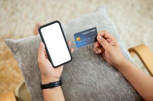 A man holding a credit card and a smartphone mockup while sitting on a lounge chair at home. photo