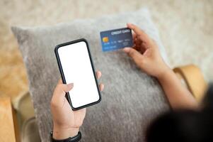 A man holding a credit card and a smartphone mockup while sitting on a lounge chair at home. photo