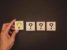Question mark and creativity concept. Hand holding a wooden cube with a drawn light bulb and a row of cubes with question marks on a black background photo