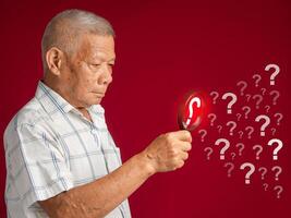 A senior man is holding a magnifying glass looking at a question mark symbol while standing on a red background photo
