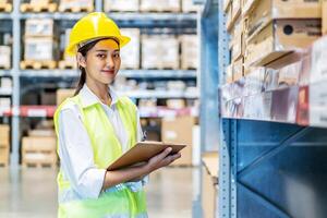 Asian woman warehouse worker in uniform doing stocktaking of products management in cardboard box on shelves in warehouse using clipboard file and pen, logistic and business export photo