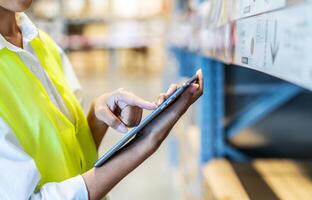 Asian manager woman doing stocktaking of products management in cardboard box on shelves in warehouse using digital tablet computer. Female professional assistant checking stock in factory. photo