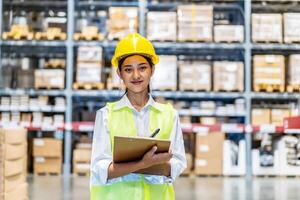 Asian woman warehouse worker in uniform doing stocktaking of products management in cardboard box on shelves in warehouse using clipboard file and pen, logistic and business export photo