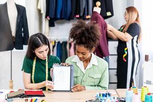 Young two fashion designers use digital tablet meeting with graphic designer discuss show sample dress design new collection on screen on desk while working in sewing workshop photo