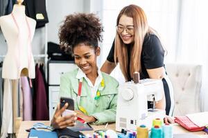 Beautiful two female african american and asian fashion designer using smartphone call to customer with happiness in tailor shop, Startup small business entrepreneur concept photo