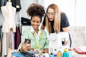 Beautiful two female african american and asian fashion designer using smartphone call to customer with happiness in tailor shop, Startup small business entrepreneur concept photo