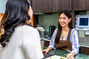 Smiling young asian waitress in a cafe with customer for service, payment or order on counter at coffee shop. Serving, employee in small business restaurant helping a girl client at checkout or work photo