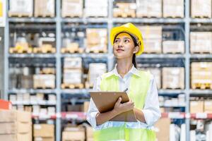Asian woman warehouse worker in uniform doing stocktaking of products management in cardboard box on shelves in warehouse using clipboard file and pen, logistic and business export photo