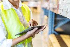Asian manager woman doing stocktaking of products management in cardboard box on shelves in warehouse using digital tablet computer. Female professional assistant checking stock in factory. photo