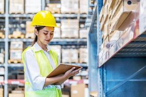 Asian woman warehouse worker in uniform doing stocktaking of products management in cardboard box on shelves in warehouse using clipboard file and pen, logistic and business export photo