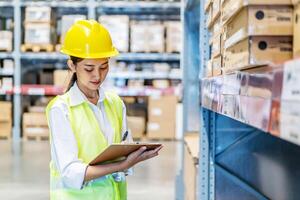 Asian woman warehouse worker in uniform doing stocktaking of products management in cardboard box on shelves in warehouse using clipboard file and pen, logistic and business export photo