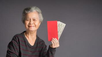 An elderly Asian woman in a sweater holding Angpao with US dollar banknote and looking at the camera with a smile while standing on gray background photo