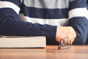 Education, knowledge, and relaxation concept. A man in a sweater with a book and glasses on a wooden table while sitting in the library. Close-up photo