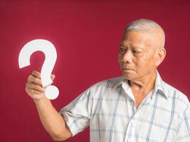 Question mark concept. Senior man holding and looking at a white question mark paper while standing with a red background in the studio. Close-up photo. Space for text photo
