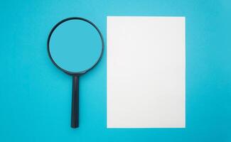 Top view of a magnifying glass and a blank white paper isolated on blue background. Search and document concept photo