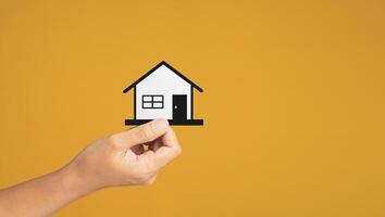 Business and real estate concept. Close-up of hand holding a mini paper house while standing on a yellow background in the studio. Space for text. Property insurance and security photo