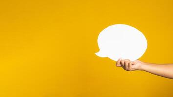 A speech bubble concept. Hand holding of an empty white speech balloon on yellow background with copy space for text. Close-up photo