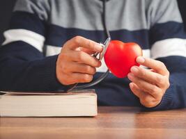 Midsection of a doctor in a sweater holding a stethoscope checking a red heart shape while sitting in the hospital or clinic. Close-up photo. Space for text. Medical, treatment, and healthcare concept photo