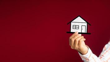 Business and real estate concept. Close-up of hand holding a mini paper house while standing on a red background in the studio. Space for text. Property insurance and security photo