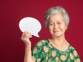 Concept of a speech bubble. Happy senior woman with short gray hair holding a blank paper and looking at the camera with a smile while standing on a red background in the studio photo
