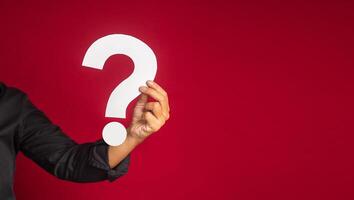 Question mark concept. Hand of a businessman in a black shirt holding a white question mark while standing with a red background in the studio. Close-up photo. Space for text photo