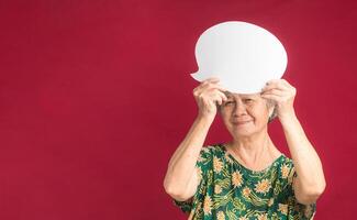 A speech bubble concept. Happy senior woman with short gray hair holding a blank paper bubble balloon and looking at the camera with a smile while standing on a red background in the studio photo
