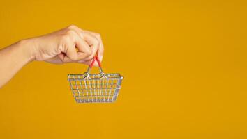 Business and e-commerce concept. Close-up of hand holding an empty mini shopping cart over a yellow background. Side view. Space for text photo